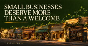 Illustrated storefront scene with the headline "Small Businesses Deserve More Than a Welcome" over a warm, lantern-lit row of local shops, TownVines.com